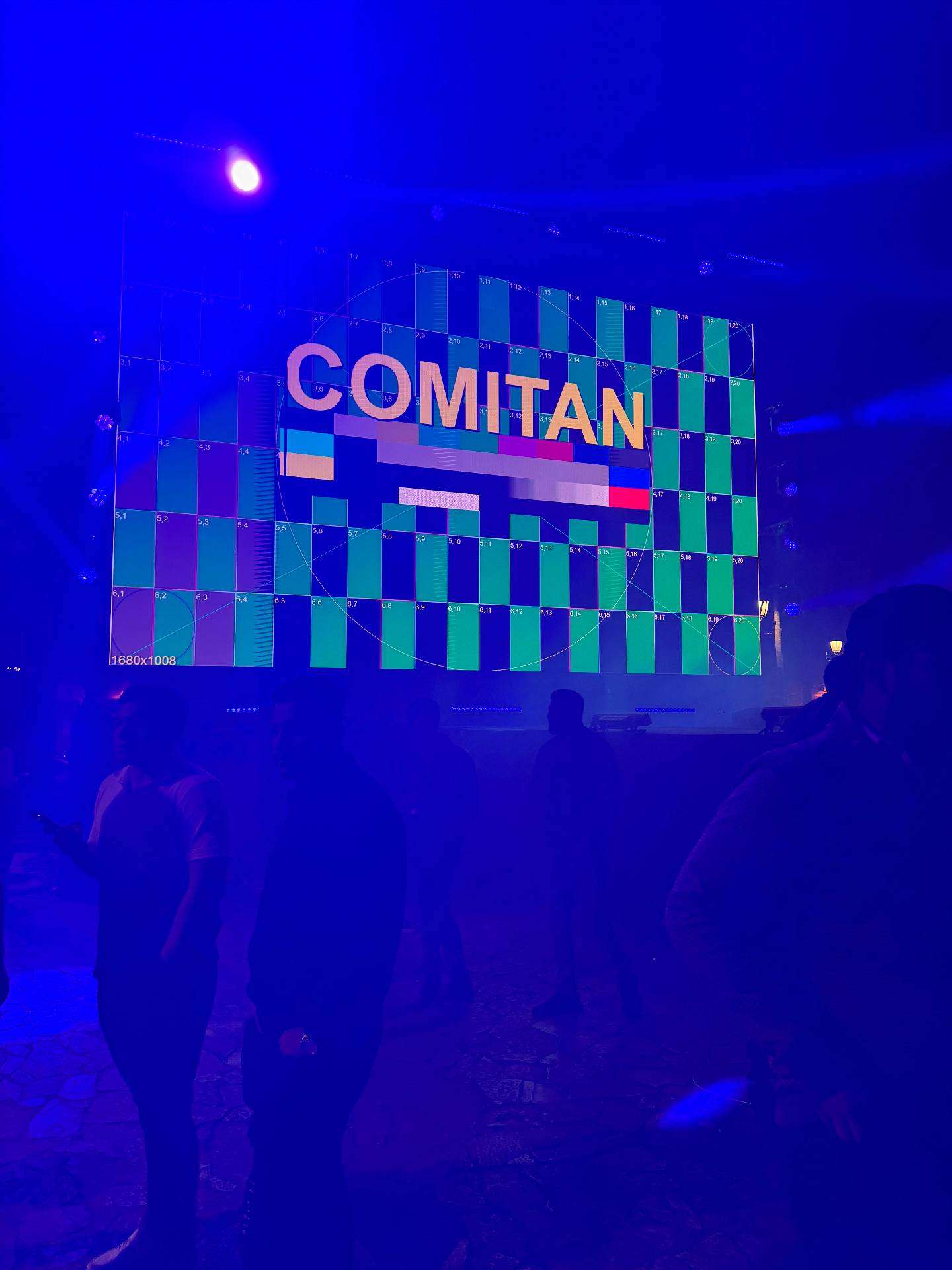 The COMITAN title on the giant event screen in blue haze and stage light.
